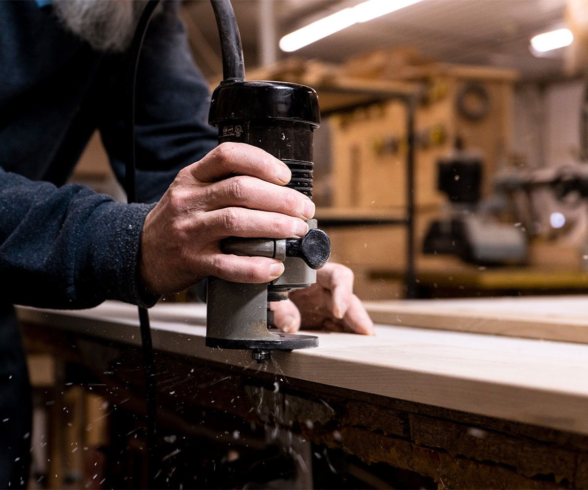 Close-up of Amish craftsman using a handheld router on a solid wood board in workshop.