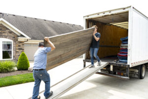 Delivery crew unloads large solid wood furniture panel from box truck down ramp to driveway.