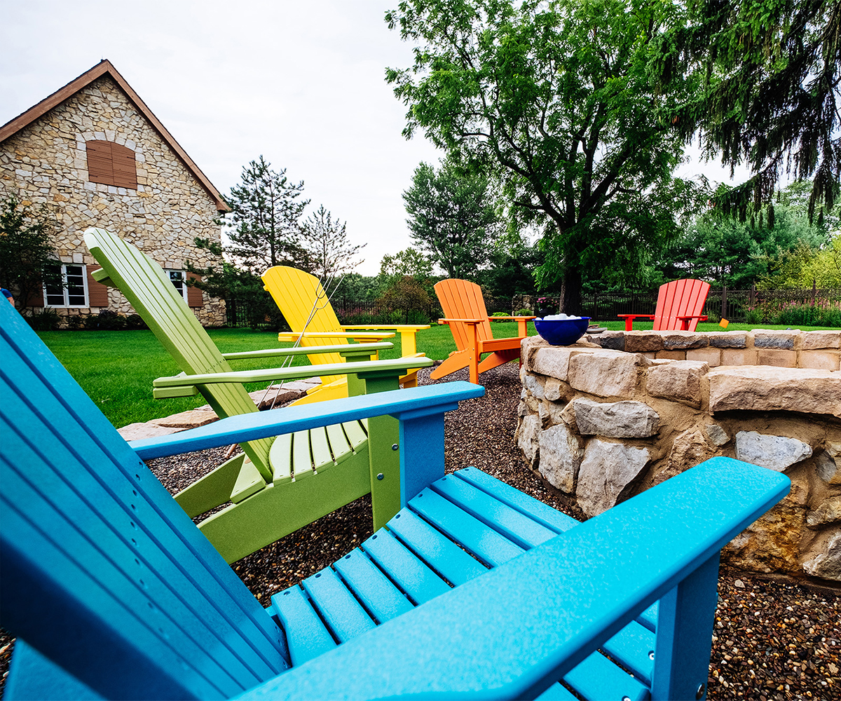 Colorful poly Adirondack chairs around a stone fire pit on lawn by farmhouse.