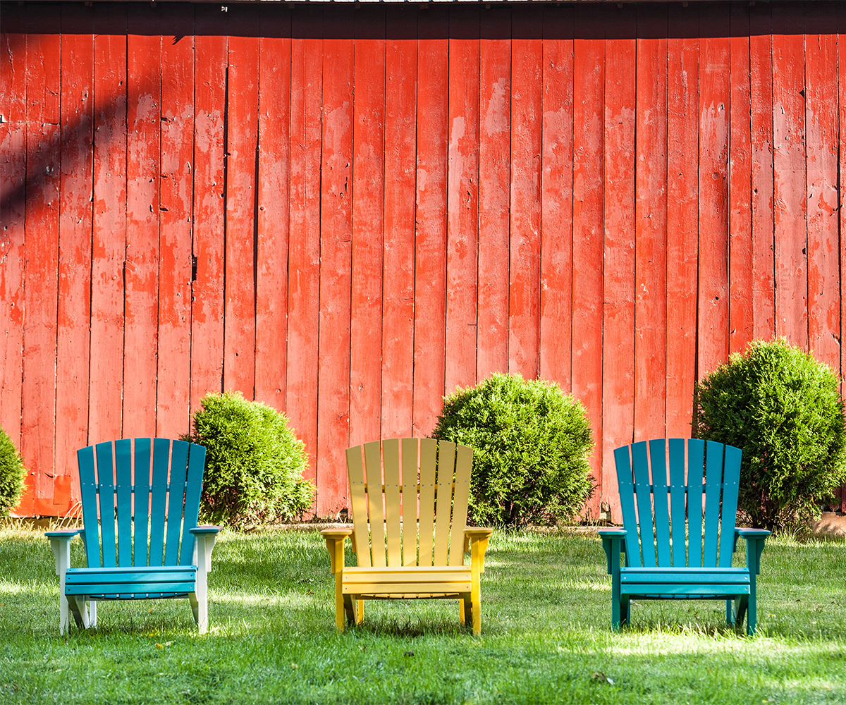 Three colorful poly Adirondack chairs before red barn and shrubs on lawn.