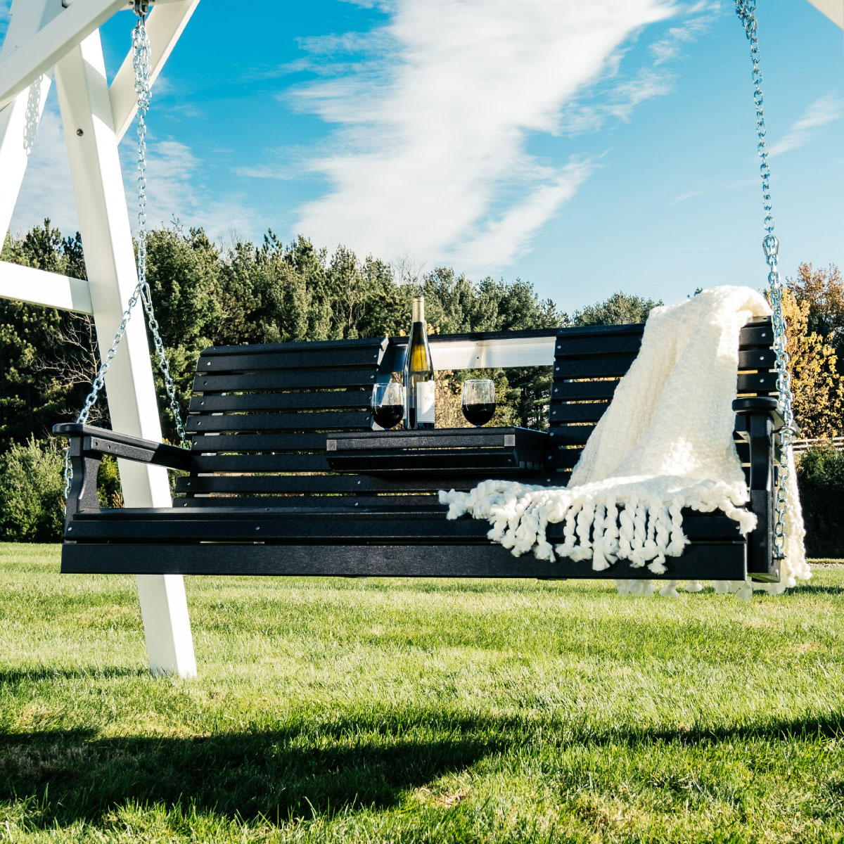 Black poly porch swing with cupholder, wine glasses, and cream throw on lawn.